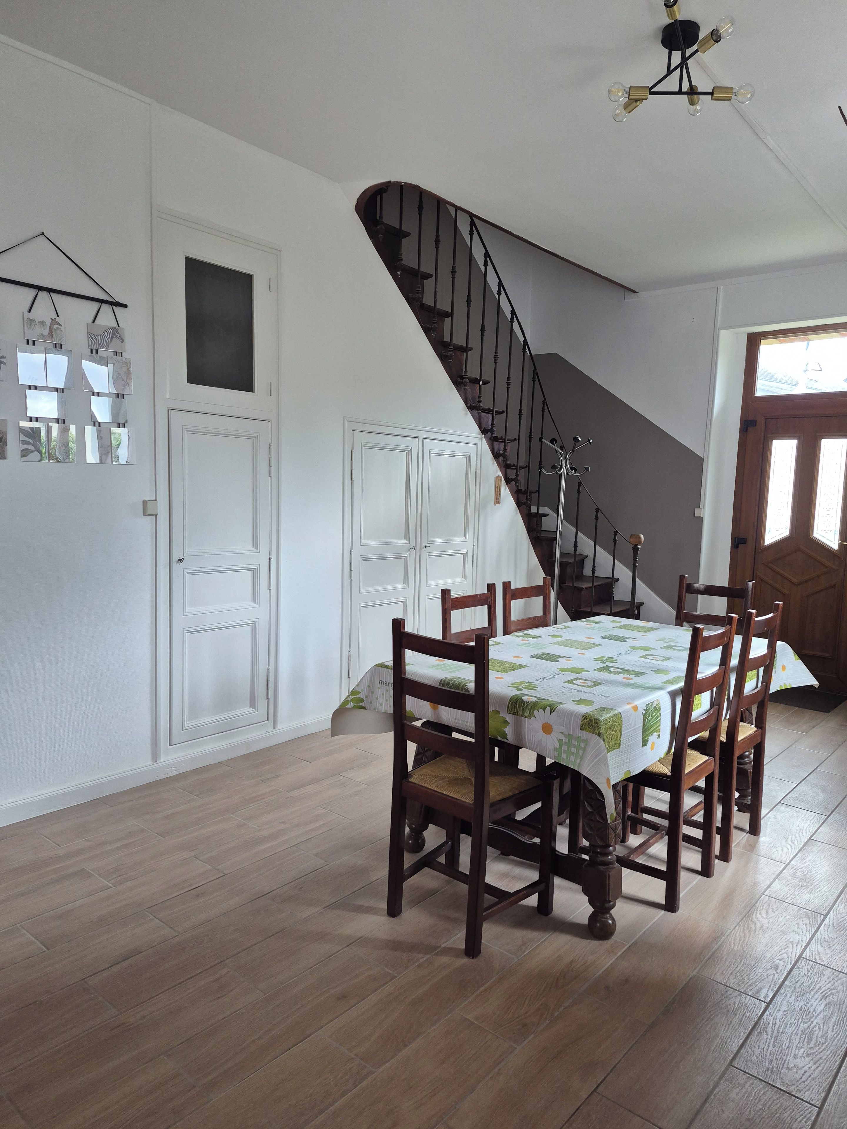 Un coin repas avec une table dressée pour quatre personnes, des chaises en bois, un escalier et un mur blanc avec une porte.