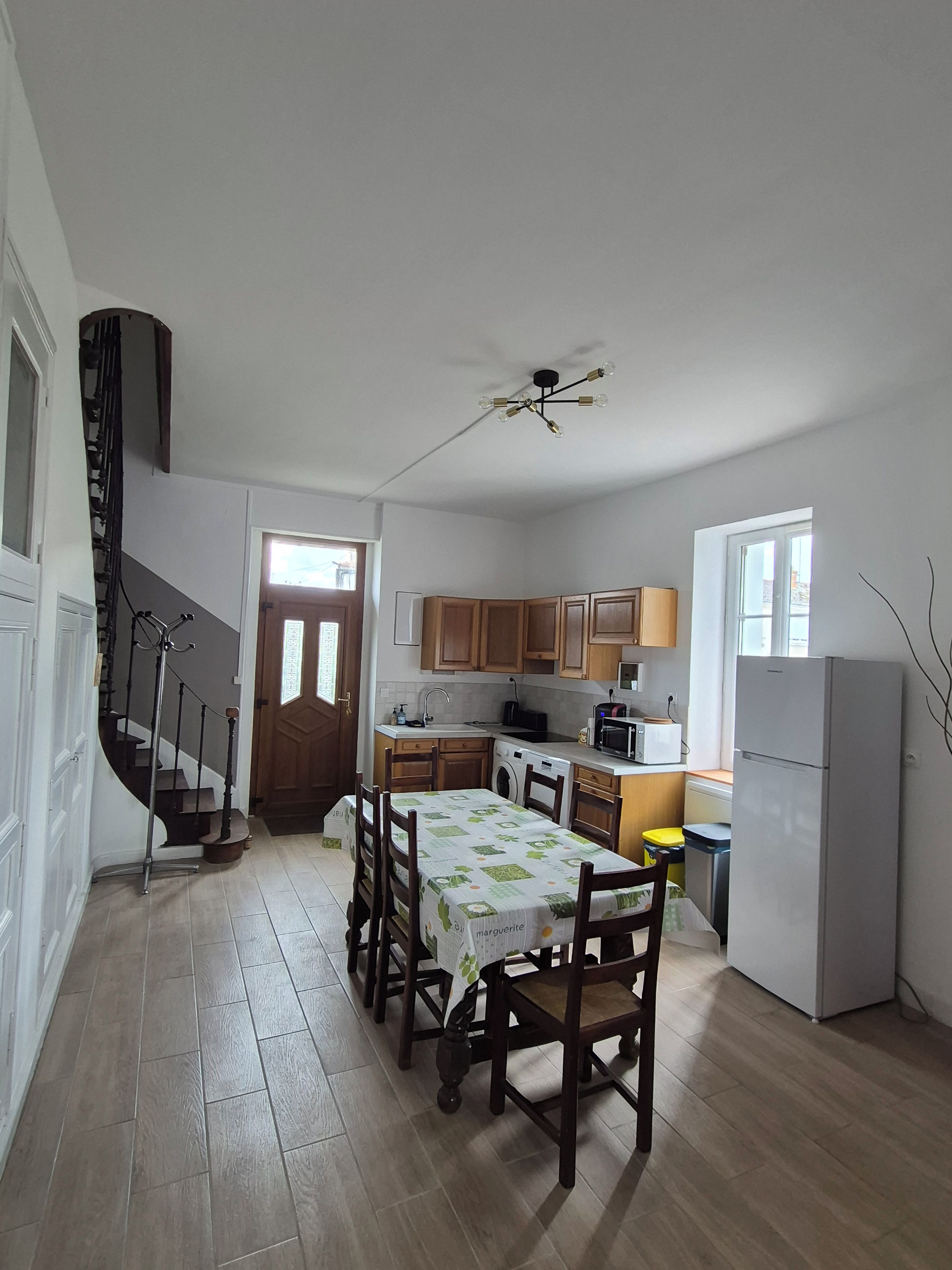 A compact kitchen with wooden cabinets, a dining table with chairs, a refrigerator, and a staircase.