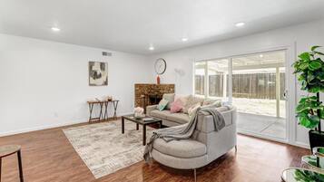 A spacious living room with hardwood flooring, a grey sofa, a coffee table, and a large window with a view of a backyard.