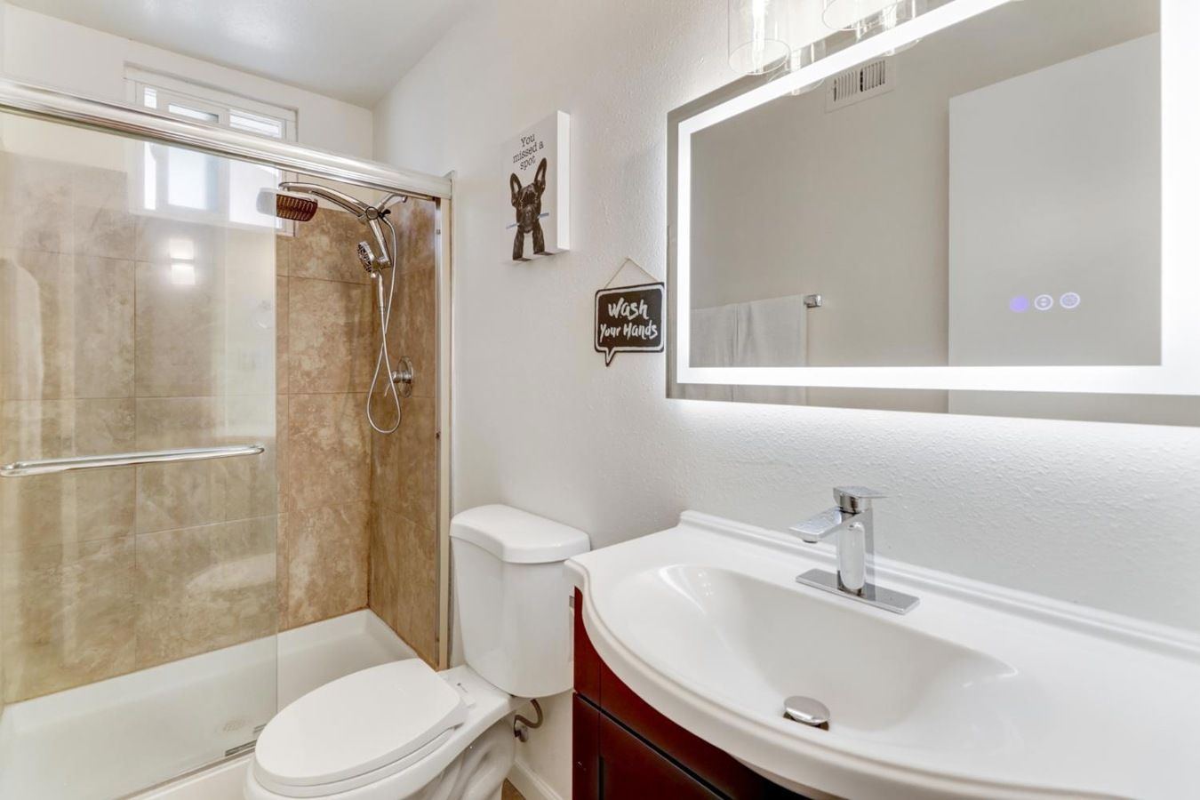 Un baño moderno con ducha envuelta en vidrio, inodoro blanco, tocador con espejo grande y dispensador de jabón de pared.