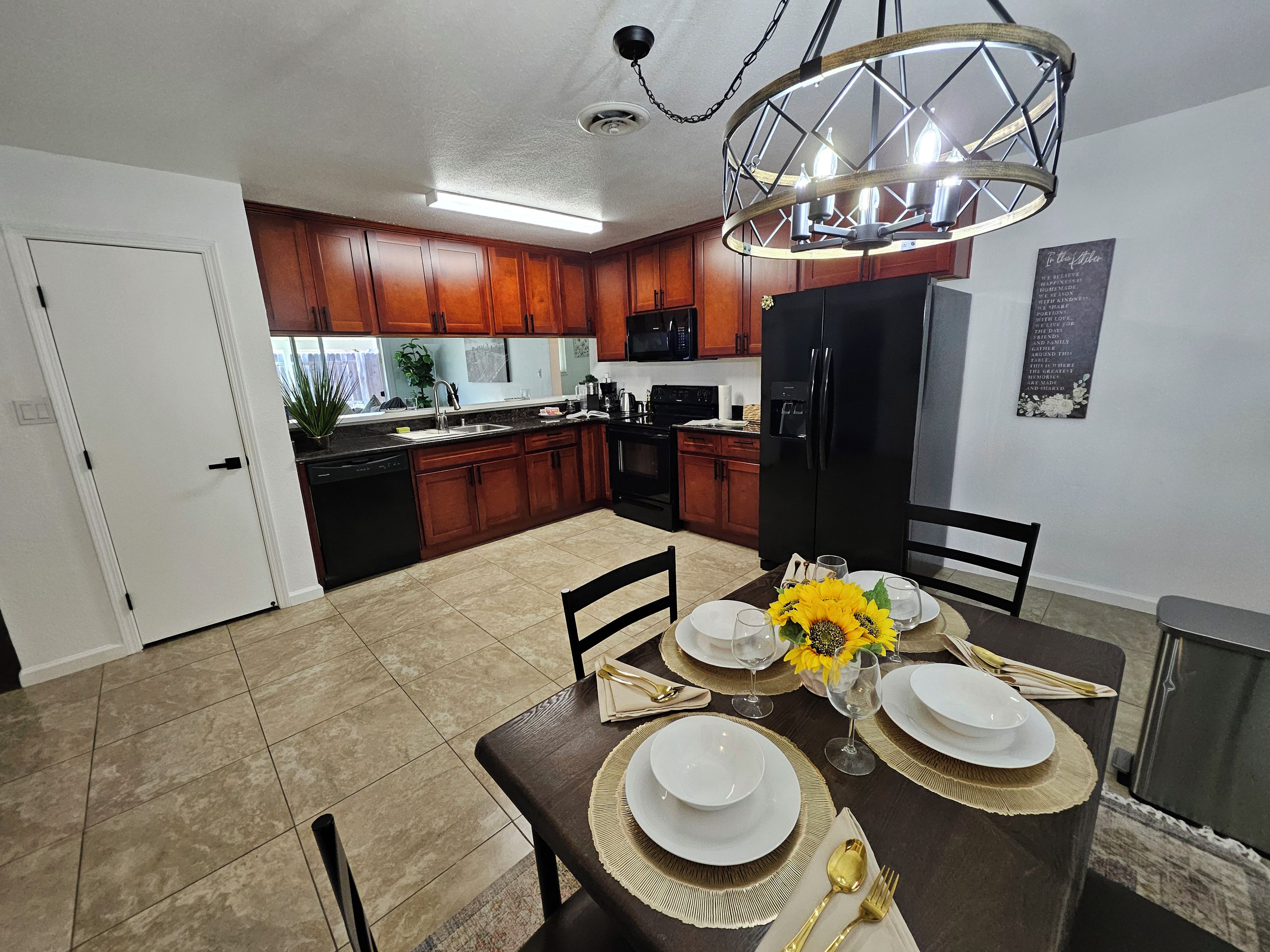 Une cuisine moderne avec des meubles en bois, un réfrigérateur noir et un coin repas aménagé pour quatre personnes, avec une table, des chaises et un vase rempli de tournesols.