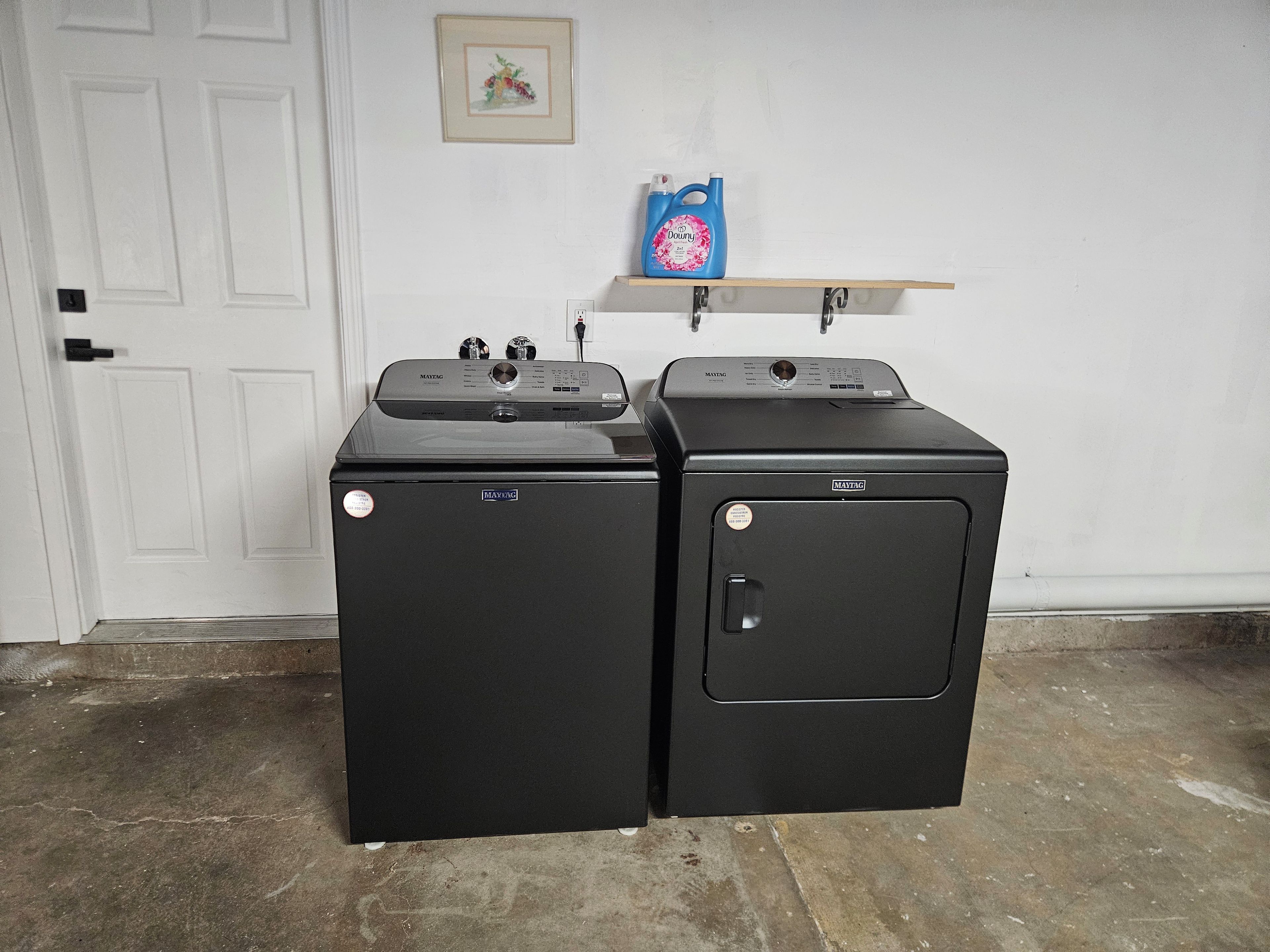 Twee zwarte Maytag wasmachines in een ruimte met een witte deur, een lijst met een foto aan de muur, en een plank met een blauwe fles wasmiddel.