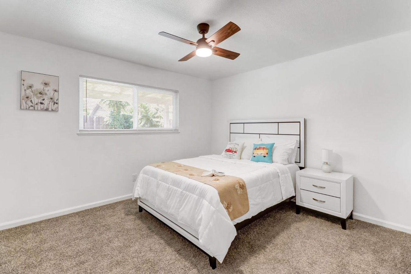 A bedroom with a bed, a nightstand, a ceiling fan, a window with blinds, and a wall-mounted artwork.