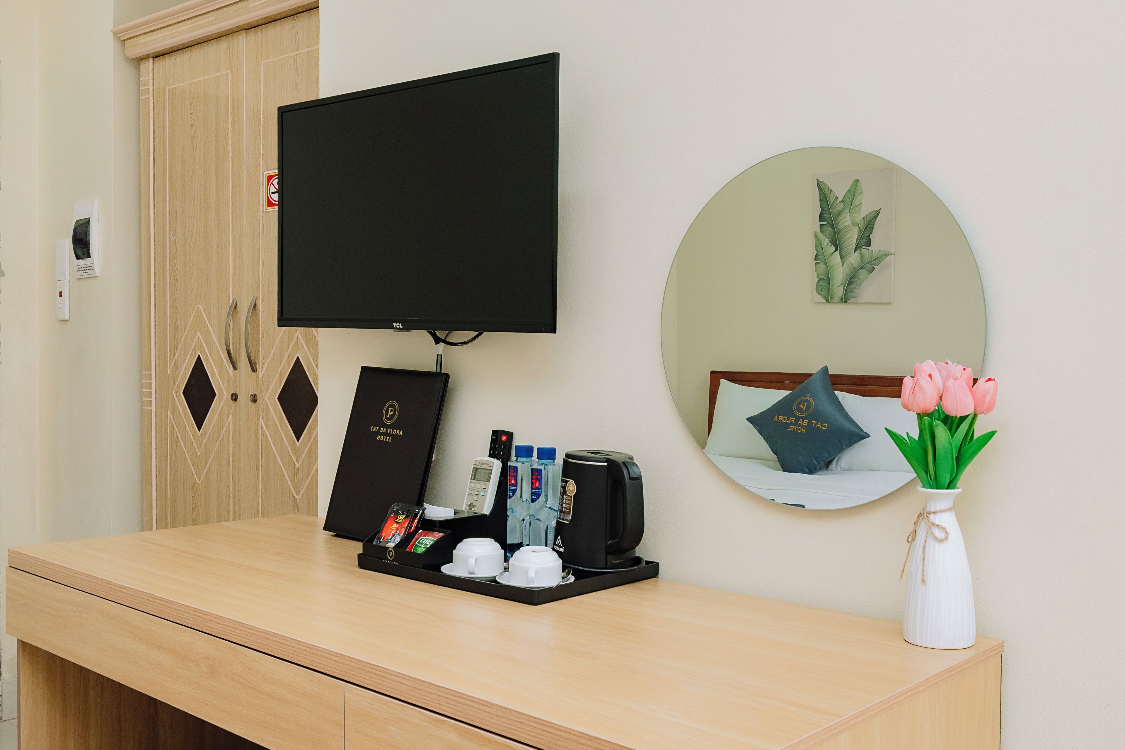 En hotelfriktionsdisk med et fladskærms-tv, et spejl, en vase med blomster og en bakke med faciliteter, herunder en kaffemaskine, vandflasker og en fjernbetjening.