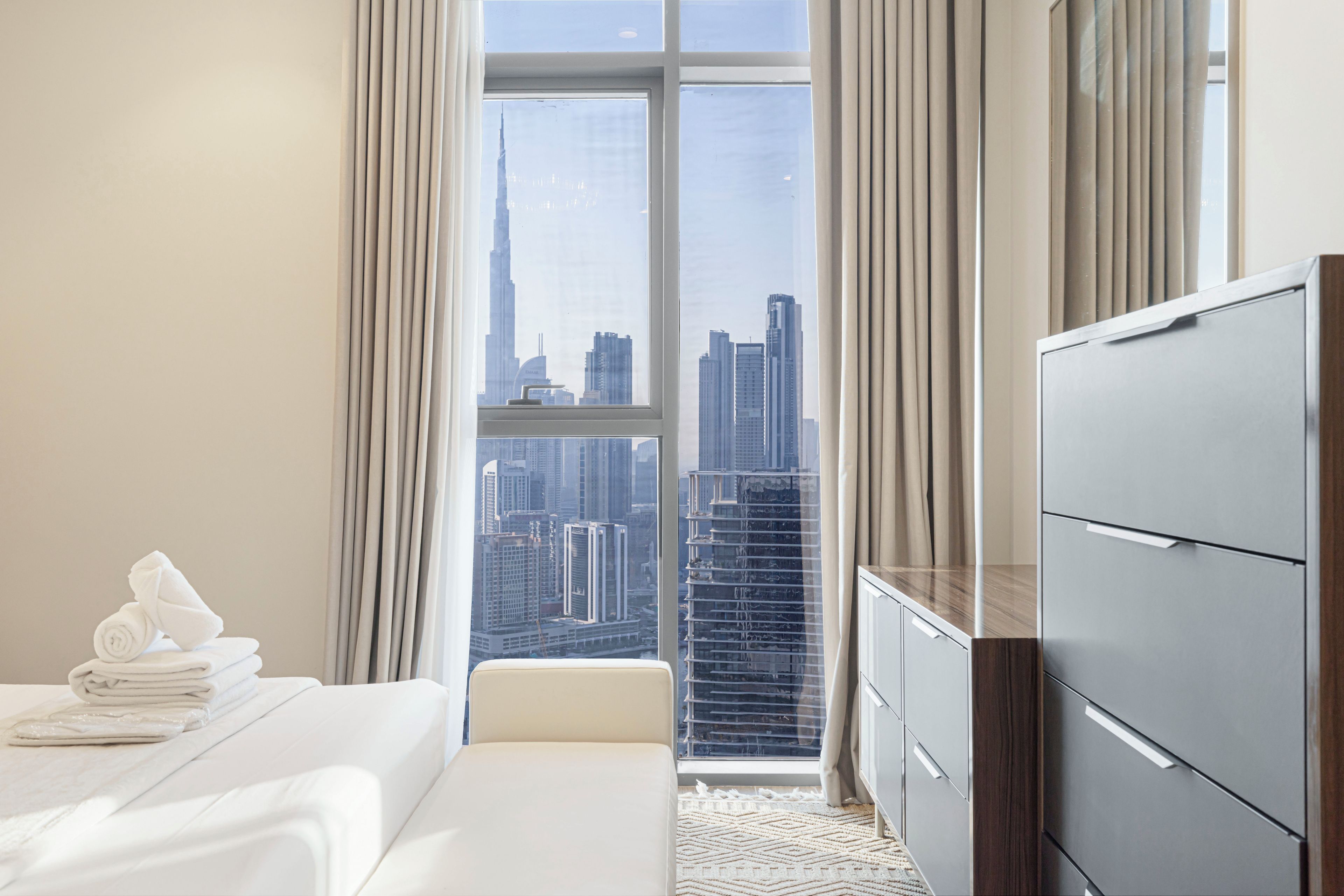 Quarto de hotel com cama, penteadeira e vista para a paisagem urbana através de grandes janelas.