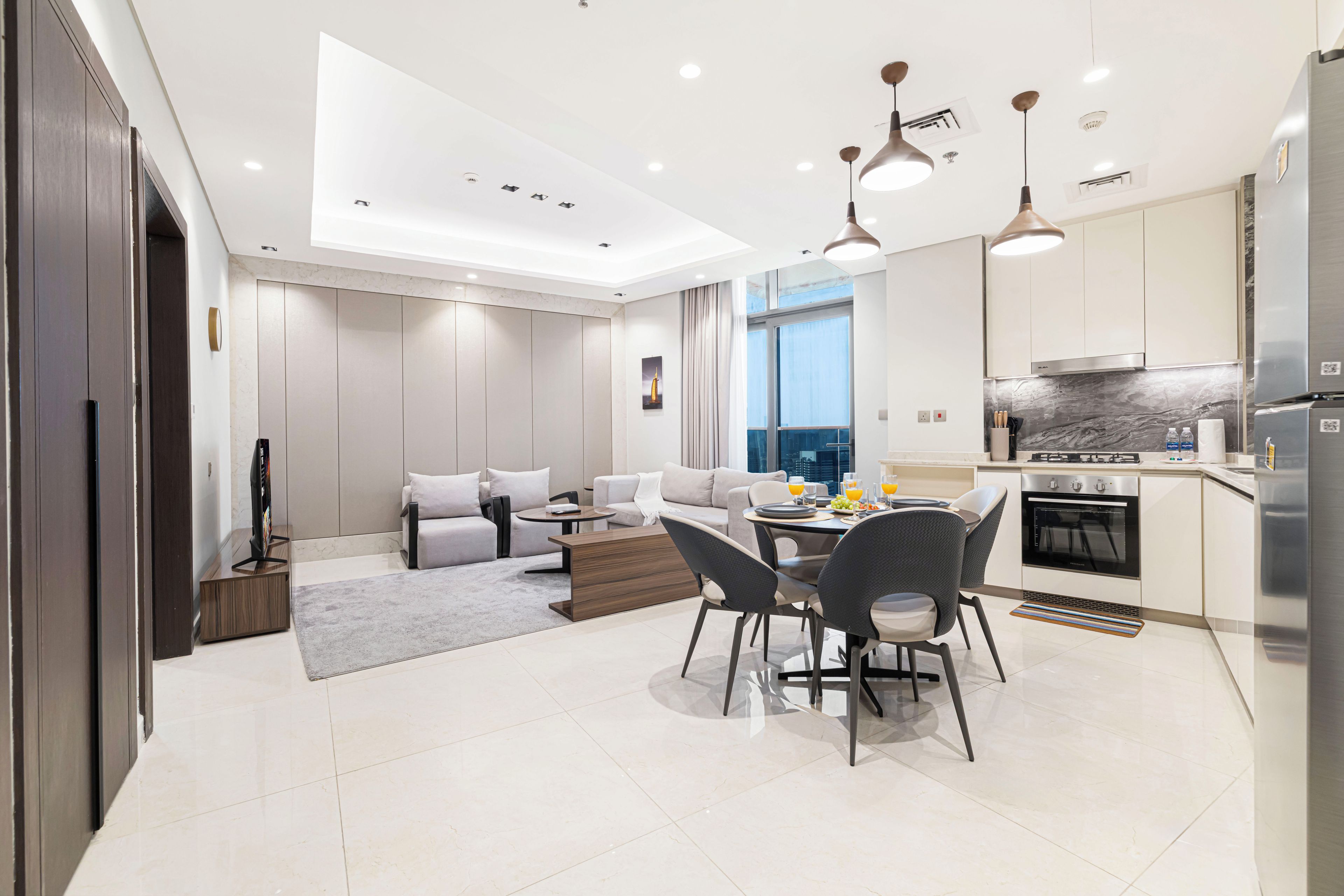 Uma sala de estar moderna com sofá, mesa de jantar e cadeiras. Há uma cozinha equipada com geladeira, fogão e pia.