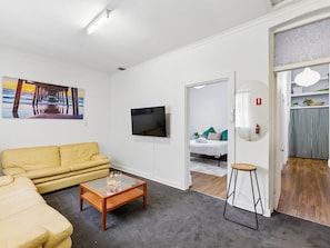 A living room with a yellow sofa, a coffee table, a flat-screen TV, and a painting of a pier.