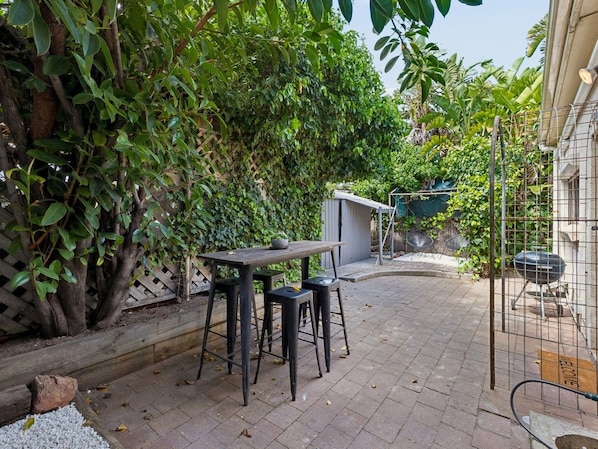 A patio area with a table and stools, surrounded by dense foliage and a small shed.