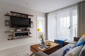 Ein modernes Wohnzimmer mit einem Flachbildfernseher, Bücherregalen, einem Couchtisch mit Zeitschriften, einem Sofa und einem großen Fenster mit transparenten Vorhängen.