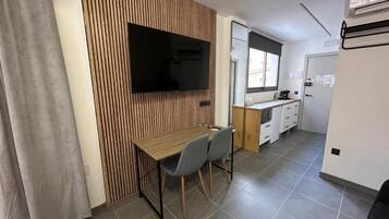 Ein kompakter Raum mit einer Wand im Holzdesign, einem Flachbildfernseher, einem kleinen Esstisch mit Stühlen, einer Küchenzeile und einem Fenster mit Jalousien.
