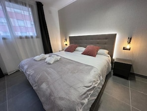 A neatly made bed with pillows and towels, a bedside table with a lamp, and a window with curtains.