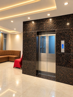 Ein moderner Aufzug mit digitalem Display, ein roter Sessel und ein braunes Ecksofa in einer Lobby mit einer Wand aus schwarzen und goldenen Flecken.