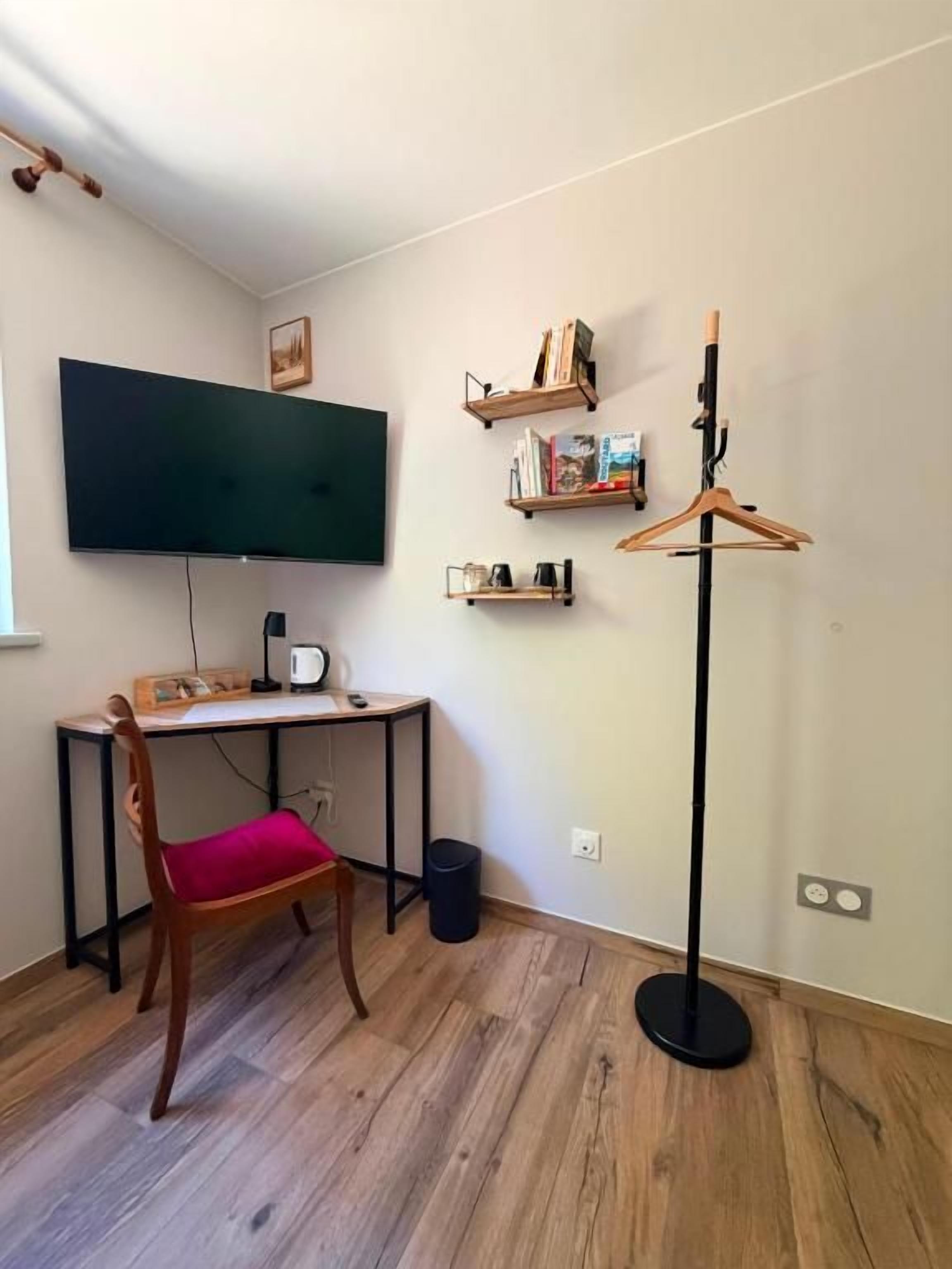 Una habitación con una televisión de pantalla plana montada en la pared, un escritorio con una computadora, una silla, una lámpara de pie y una estantería.
