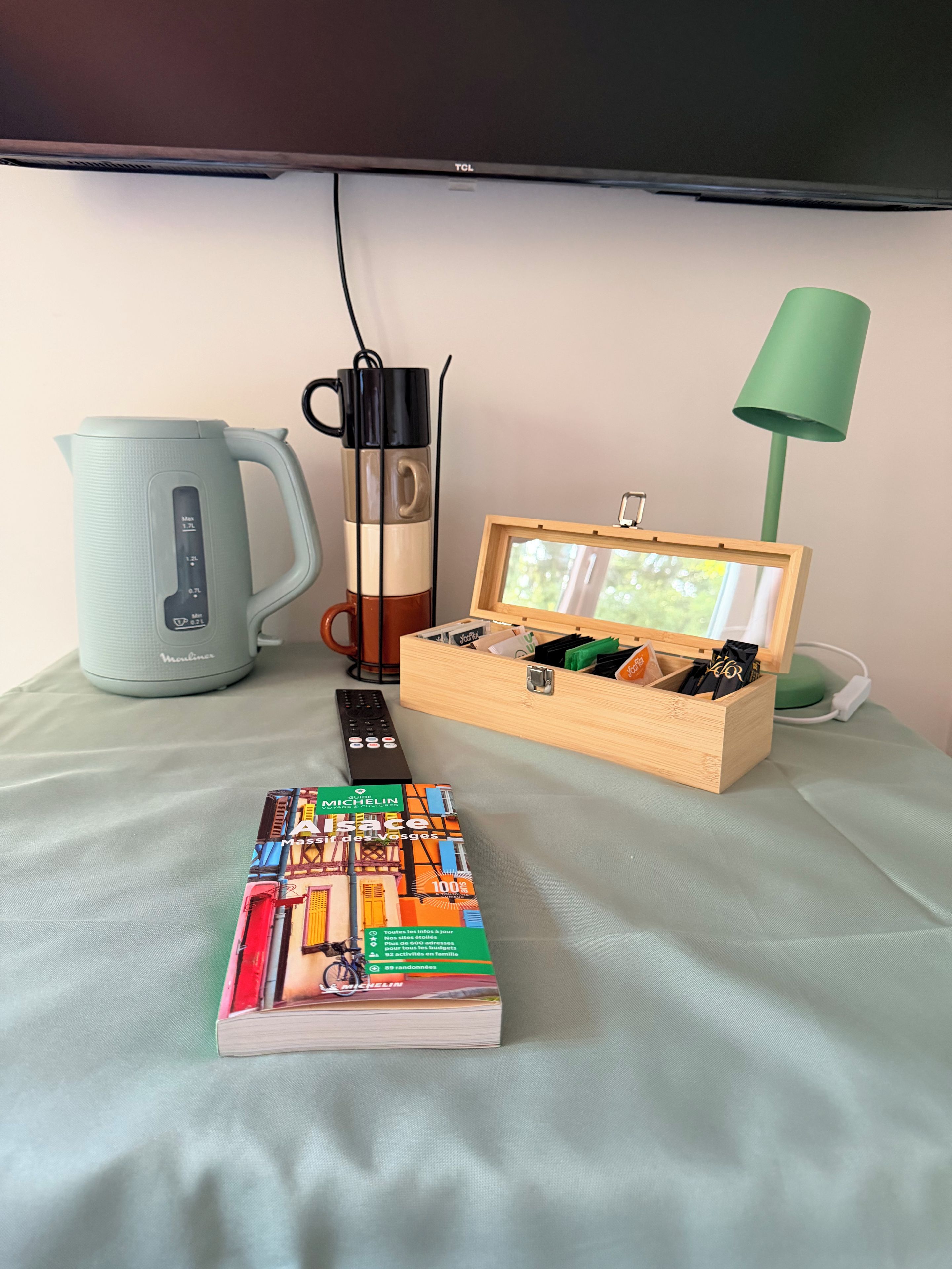 Een kamer met een bed, nachtkastje, lamp, waterkoker, mok, afstandsbediening en een boek.