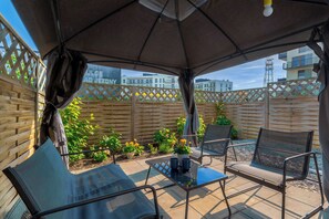 En udendørs patio med bord og stole, et træhegn og udsigt til bygninger i baggrunden.