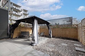 A covered outdoor seating area with a patio table and chairs, surrounded by a wooden fence and a building with balconies. - Kolobrzeg | Bałtycka 11B | Cosy Apartment | Private garden (Kołobrzeg)