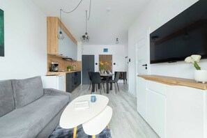 A modern living space with a sofa, a dining table with chairs, a kitchen area, and a flat-screen TV mounted on the wall. - Kolobrzeg | Bałtycka 11B | Cosy Apartment | Private garden (Kołobrzeg)