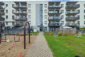 A multi-story residential building with balconies, a playground with swings and sand, and a brick pathway. - Kolobrzeg | Bałtycka 11B | Cosy Apartment | Private garden (Kołobrzeg)