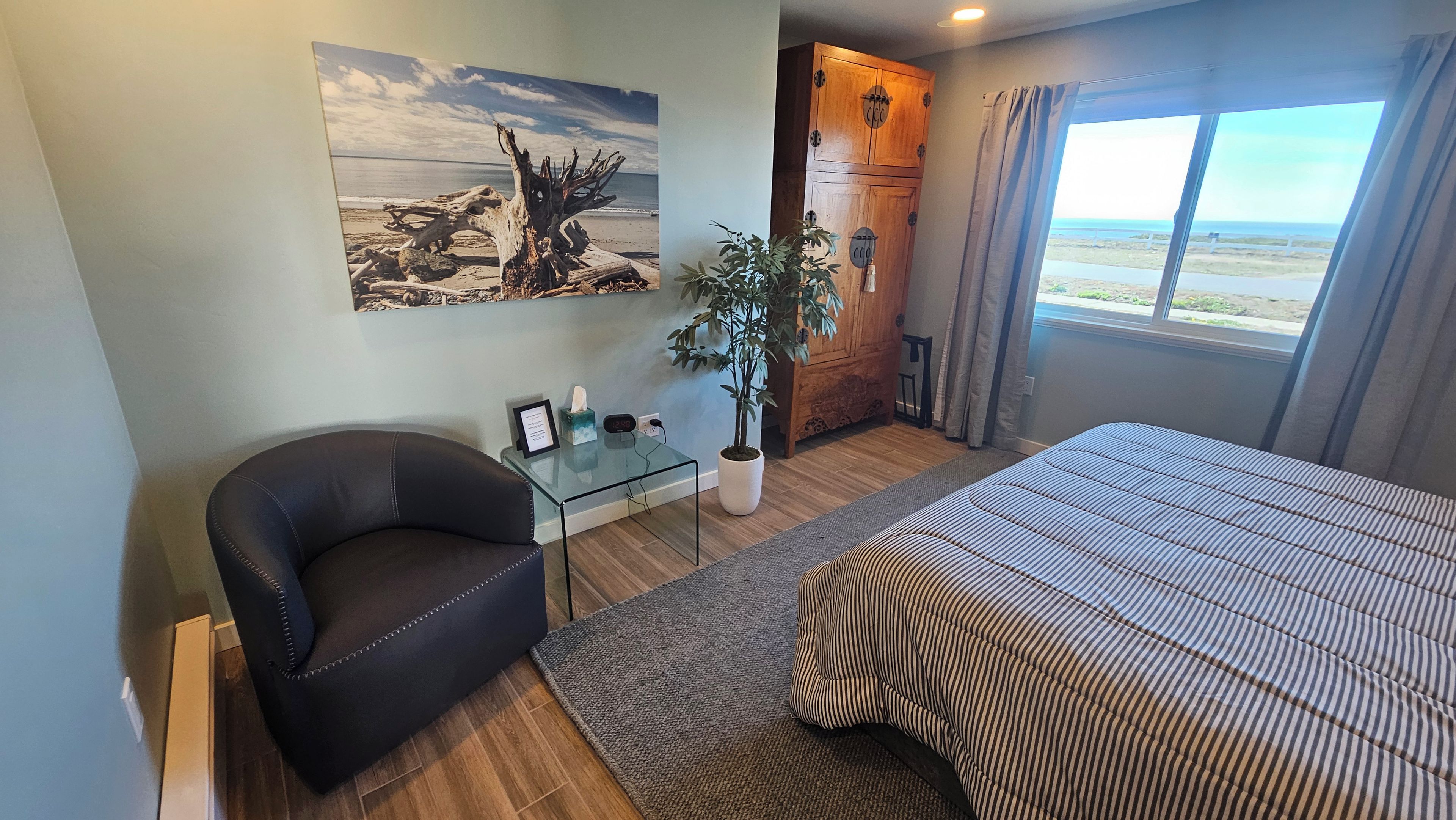 A hotel room with a bed, a chair, a desk, a potted plant, and a painting on the wall.