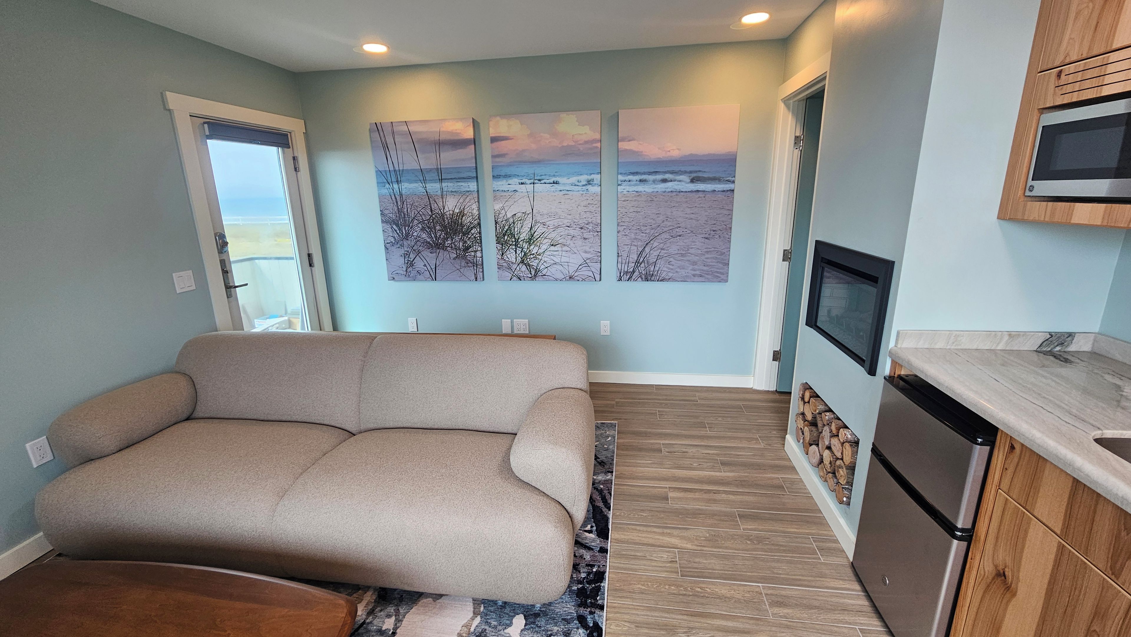 A modern living room with a sofa, a kitchen area with a microwave, and a large window with a view of the beach.
