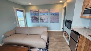 A modern living room with a sofa, a kitchen area with a microwave, and a large window with a view of the beach. - Point Arena Lighthouse - Driftwood Suite (Point Arena)