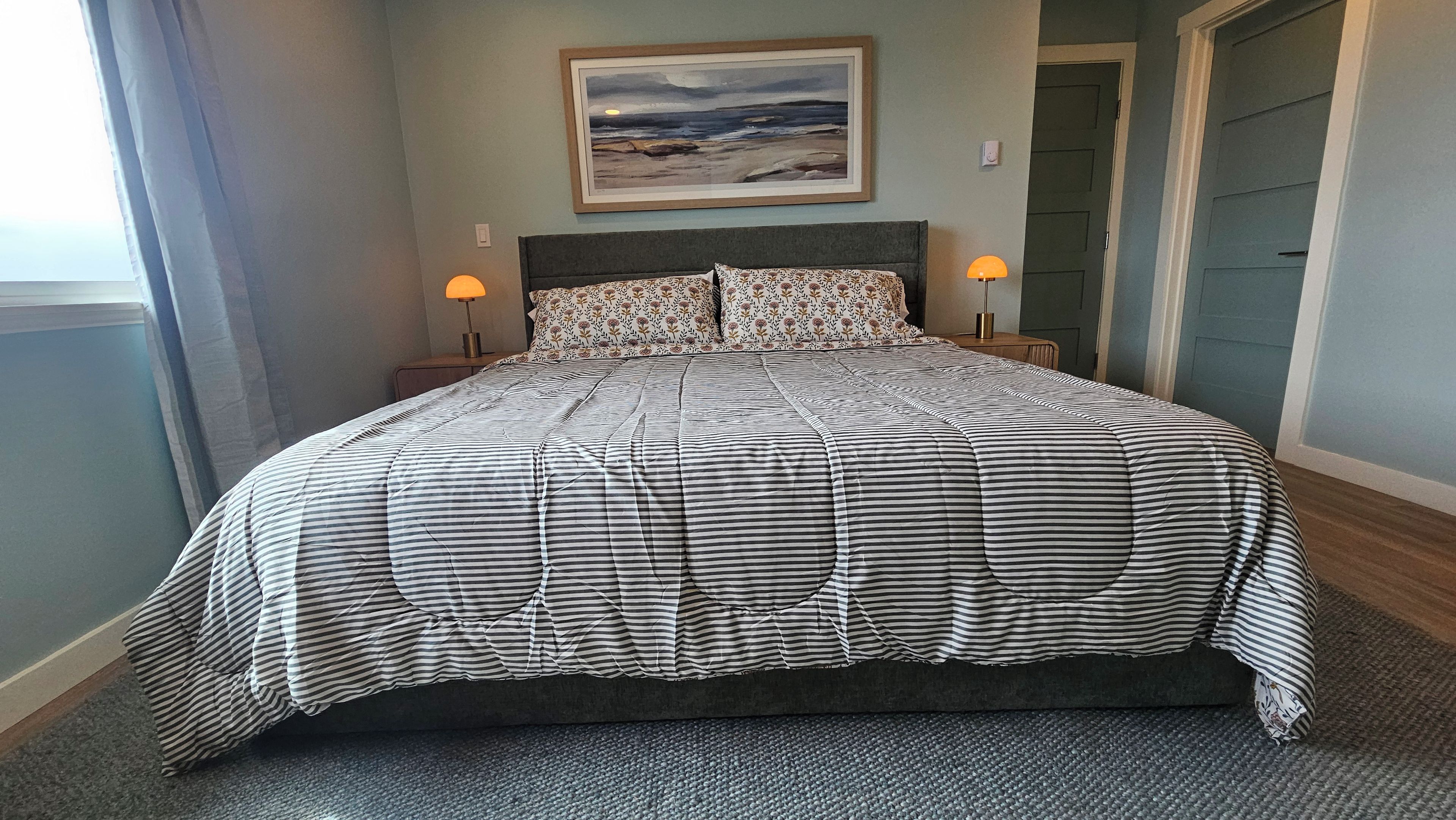 A bed with a striped bedspread, two bedside lamps, a framed picture on the wall, and a door in the background.