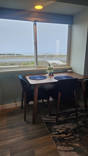 A dining area with a table and chairs, a view of the beach, and a window with blinds. - Point Arena Lighthouse - Driftwood Suite (Point Arena)