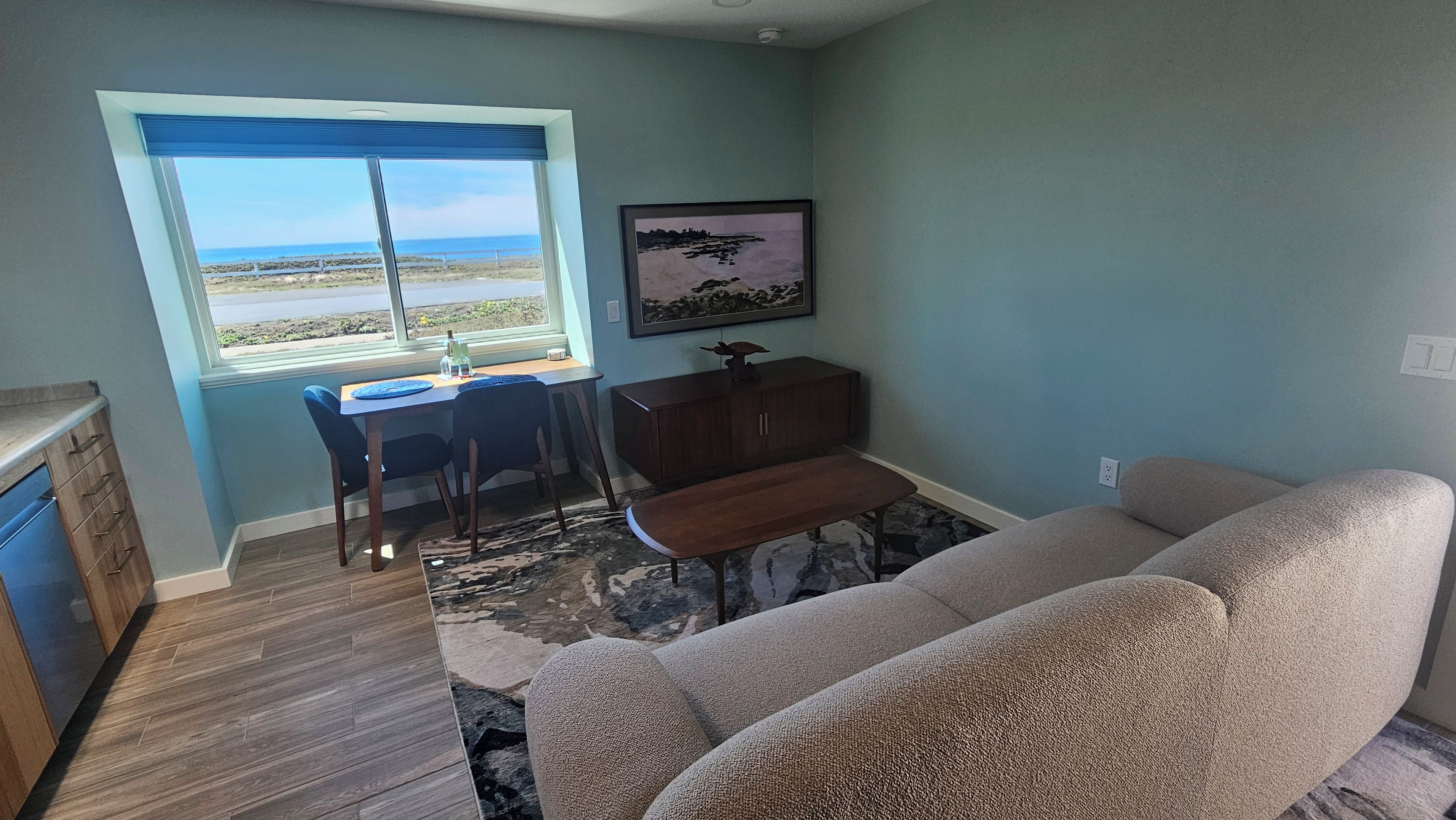 A living room with a view of the beach, a dining area with a table and chairs, and a large beige sofa.