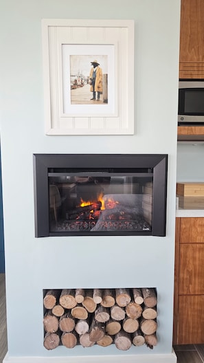 A fireplace with a framed picture above it, a modern kitchen with a microwave, and a stack of firewood below.