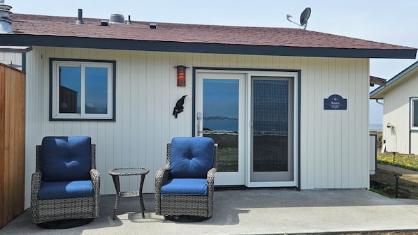 A small building with a white exterior, a red roof, and a sign that reads 'Raven Studio.' There are two wicker chairs with blue cushions and a small round table in front of the building.