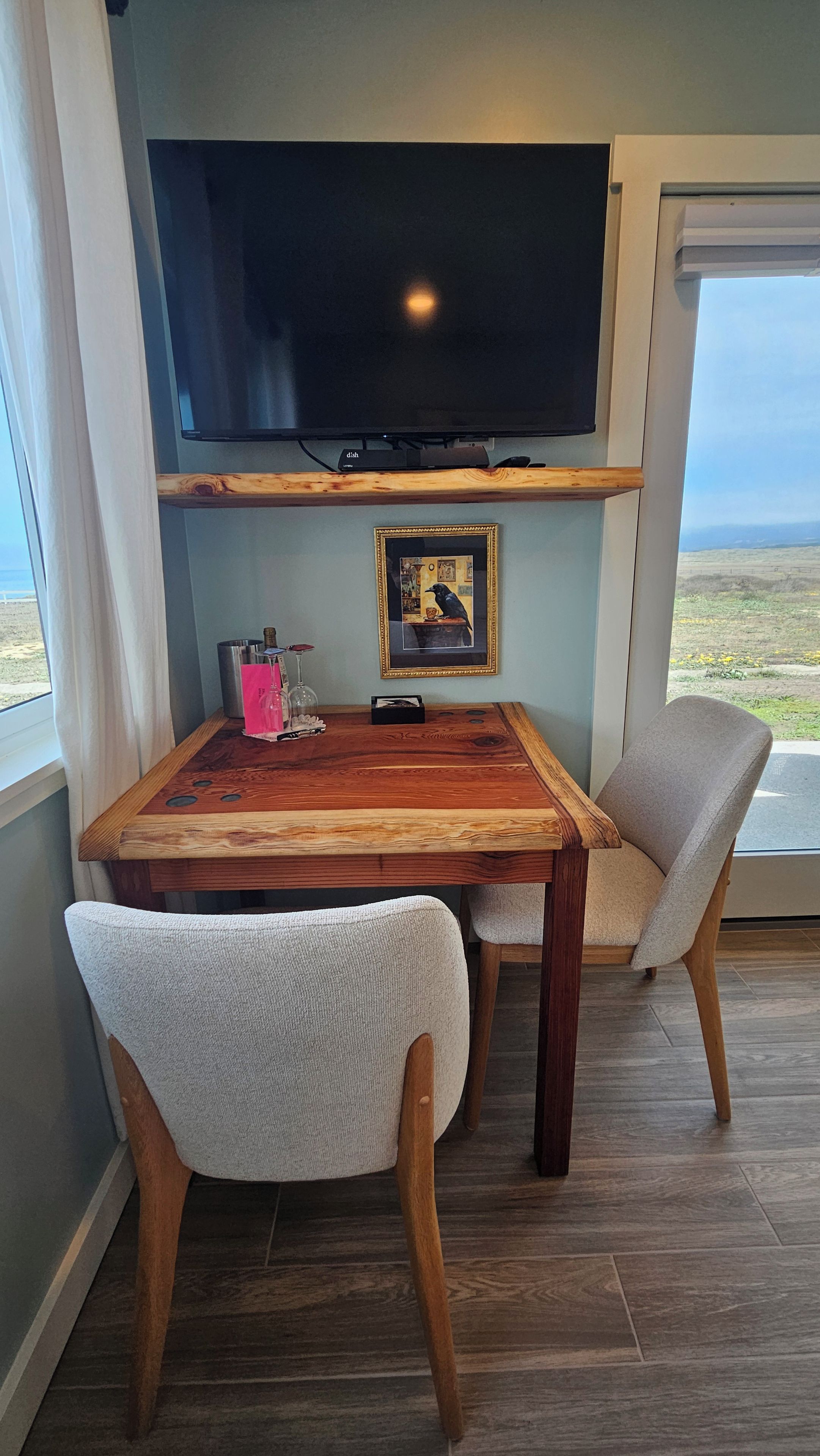 A dining area with a wooden table, two chairs, a framed picture, and a flat-screen TV mounted above.