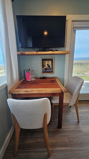 A dining area with a wooden table, two chairs, a framed picture, and a flat-screen TV mounted above. - Point Arena Lighthouse - Raven Studio (Point Arena)
