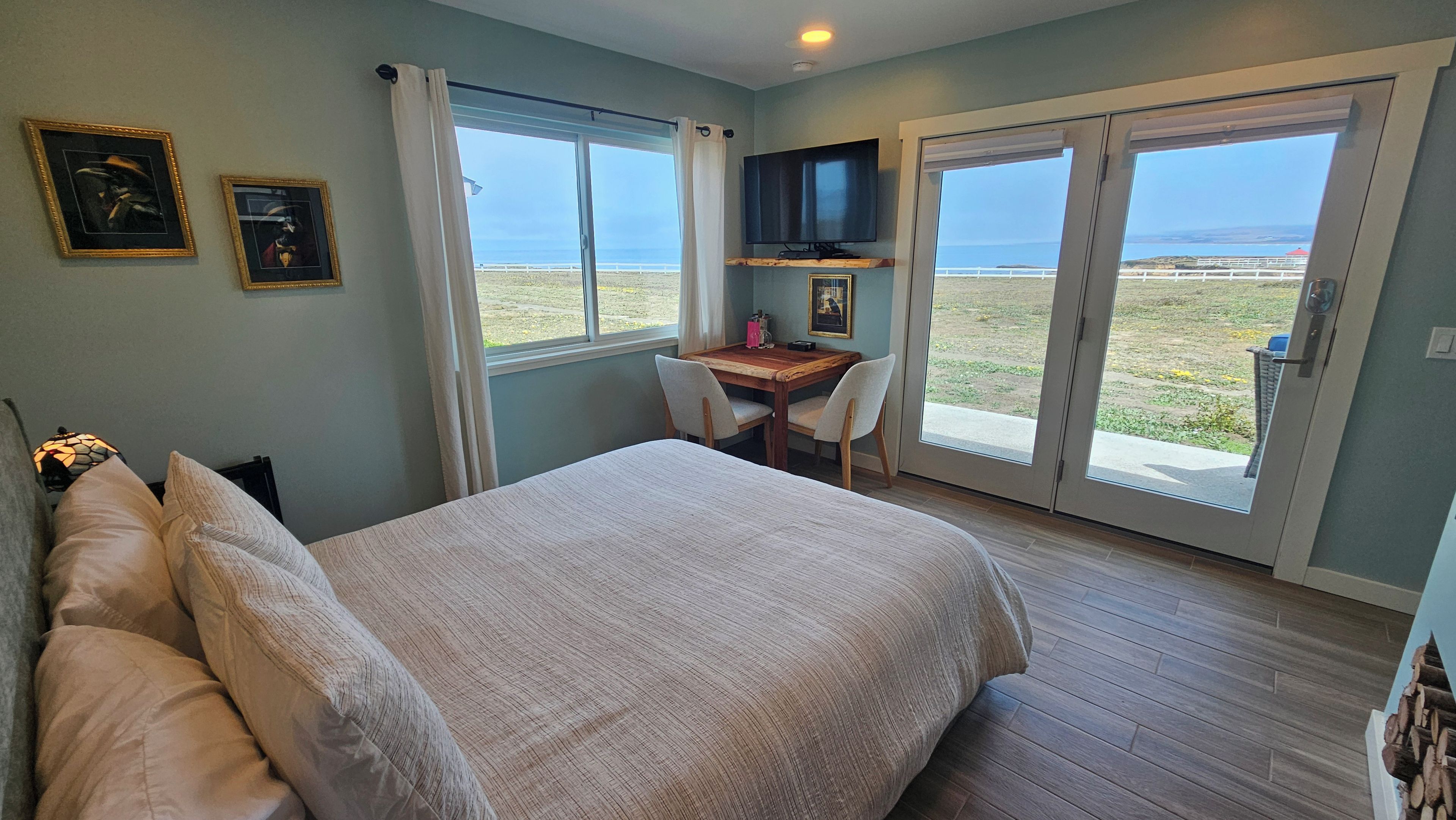 A bedroom with a bed, a desk, and a TV. There are windows with a view of the beach.