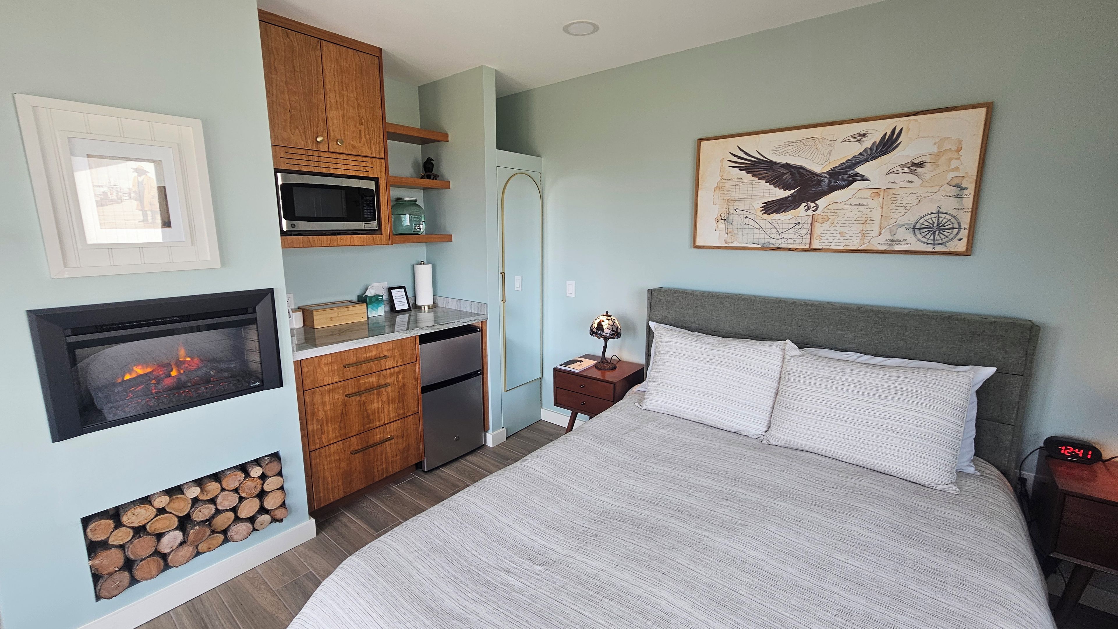 A bedroom with a bed, a nightstand, a wall-mounted lamp, a framed picture, a microwave, a refrigerator, and a fireplace.