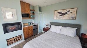 A bedroom with a bed, a nightstand, a wall-mounted lamp, a framed picture, a microwave, a refrigerator, and a fireplace.