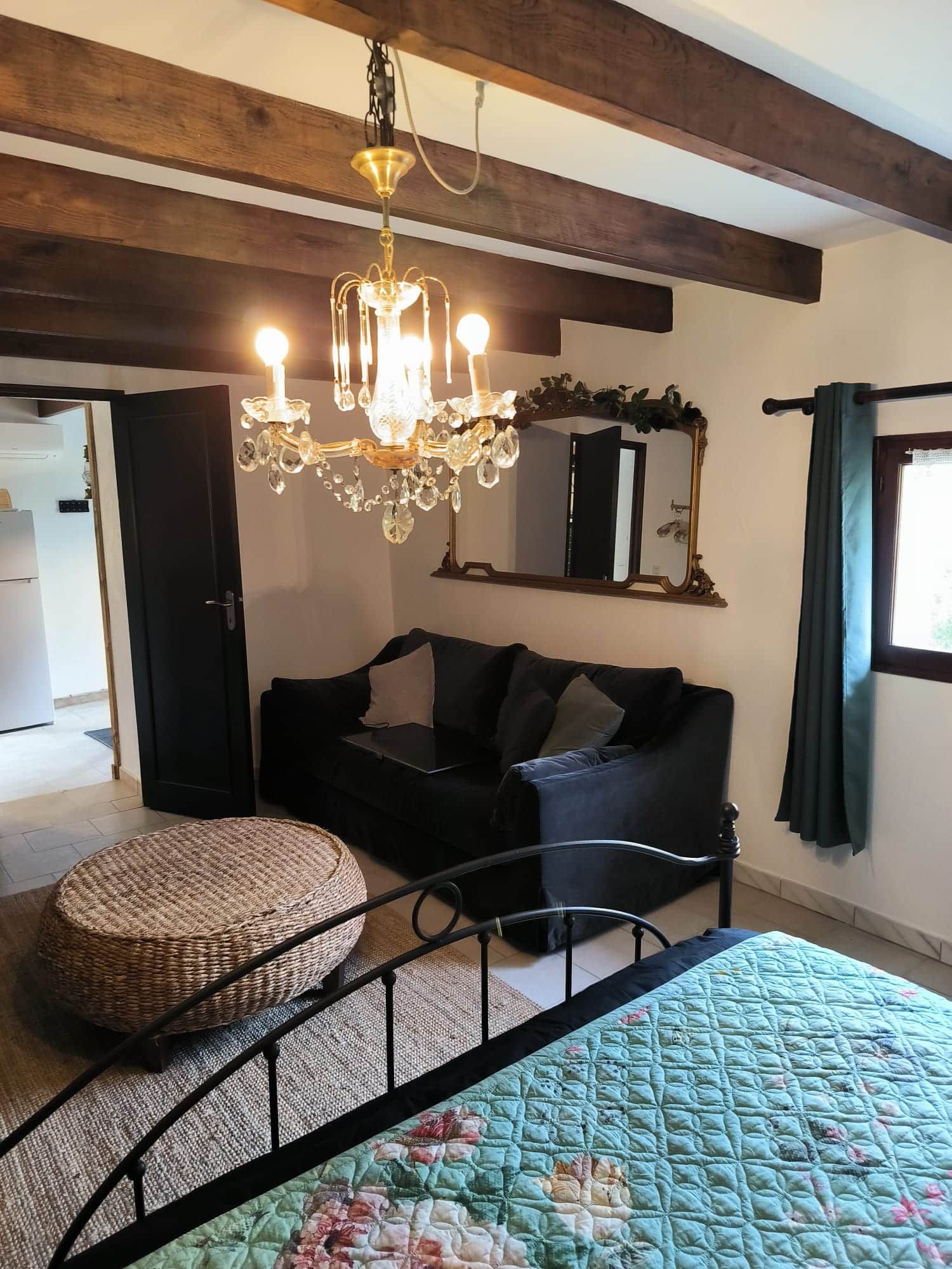 Une pièce comprenant un lit, un cadre de lit en métal noir, un pouf en osier, un canapé noir, un lustre, un miroir et une fenêtre avec des rideaux.