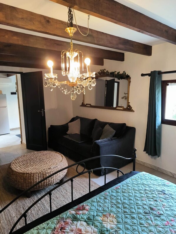 Une pièce comprenant un lit, un cadre de lit en métal noir, un pouf en osier, un canapé noir, un lustre, un miroir et une fenêtre avec des rideaux.