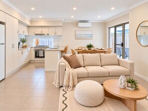 A modern kitchen with white cabinets, a dining area with a wooden table, and a beige sofa.