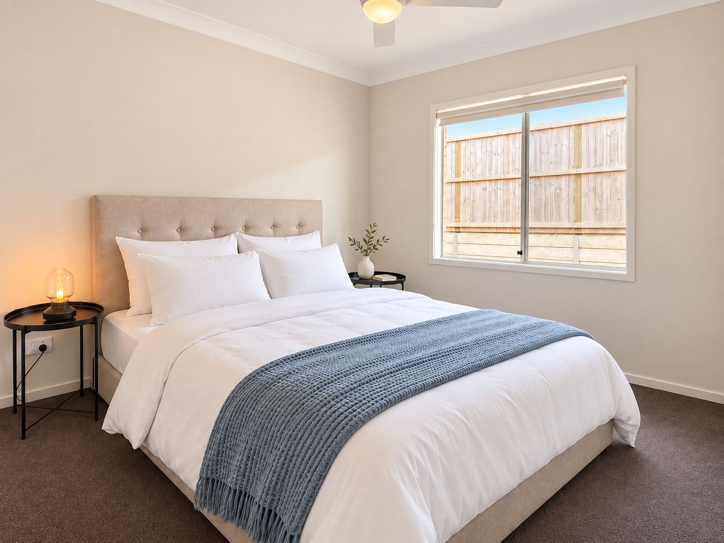 A bedroom with a bed, a nightstand, a lamp, a window with a view of a wooden fence, and a ceiling fan.