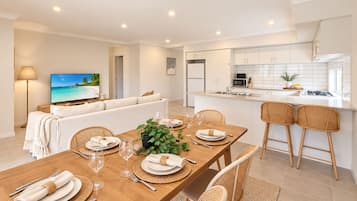 A modern kitchen with white cabinets, a dining area with a wooden table set for four, and a living room with a sofa and a TV displaying a beach scene.
