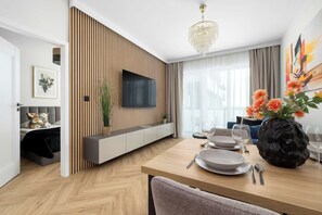 A modern living room with a flat-screen TV, wooden panel wall, and a dining area with a table set for two. - Katowice | Zabrska 15 | Modern Apartment | Air conditioning (Katowice)