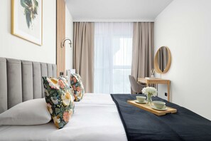 Quarto de hotel com cama, escrivaninha, cadeira, bandeja com xícara e flores, e janela com cortinas.