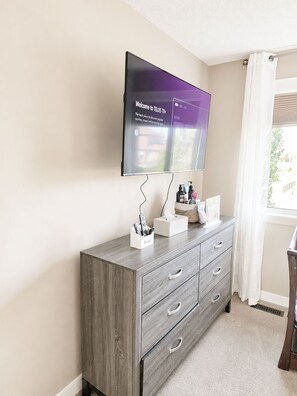 A room with a grey wooden dresser, a flat-screen TV mounted on the wall, and a window with white curtains. - Elegant and Spacious Getaway in St. Albert. 4 BR's, AC and Premium Amenities. (St. Albert)