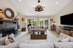 Un salon spacieux avec une cheminée en pierre, un grand canapé, une table basse agrémentée d’objets décoratifs, un téléviseur et un lustre.