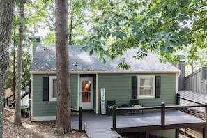 A small, single-story house with a green exterior, a front porch with a bench, and a wooden deck.