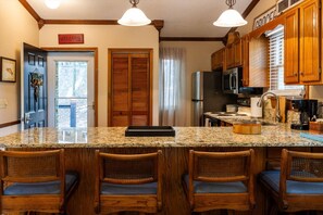 A kitchen with a granite countertop, wooden chairs, a refrigerator, and a microwave.