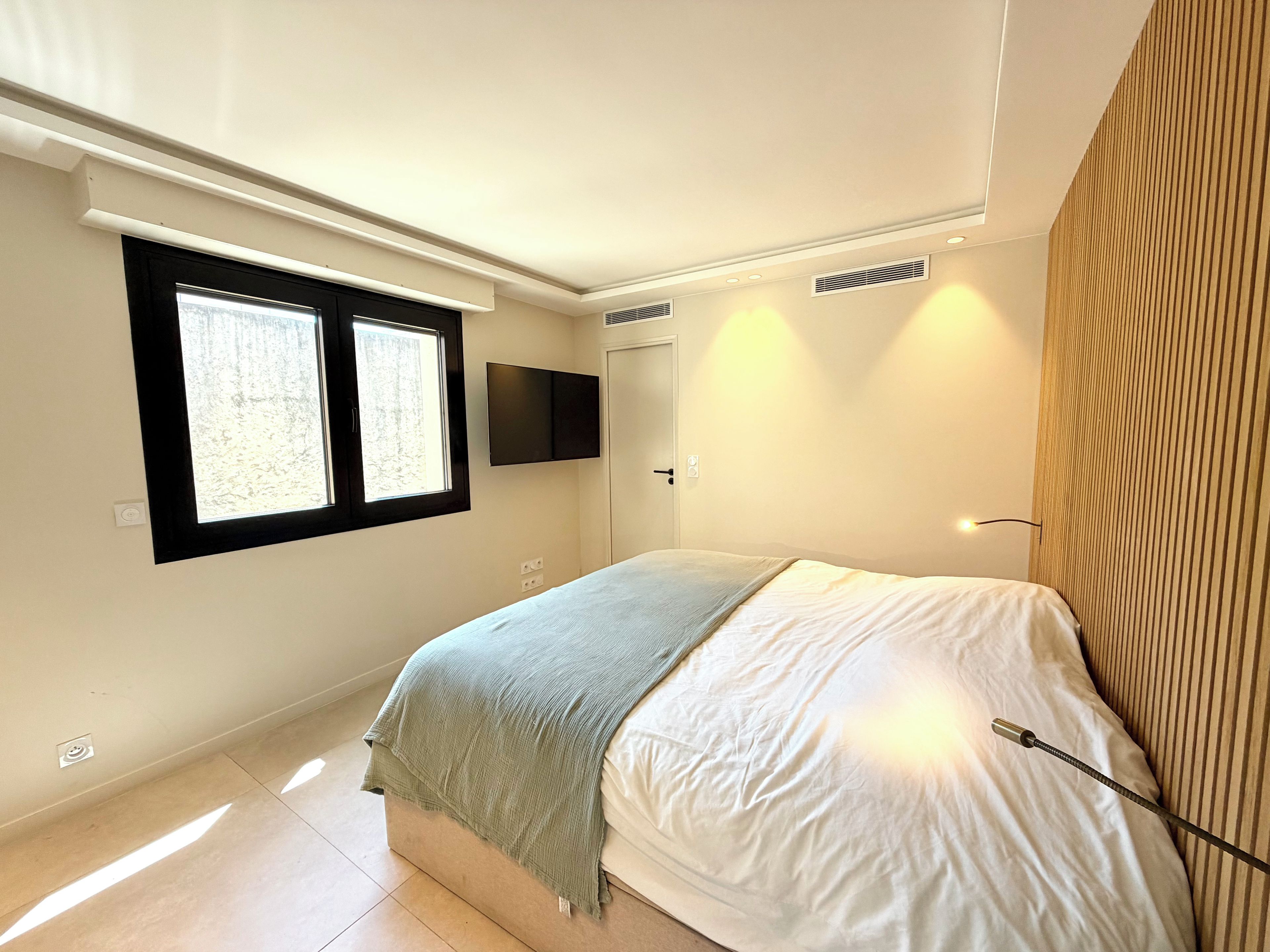 A bedroom with a bed, a window, a wall-mounted TV, and a door.