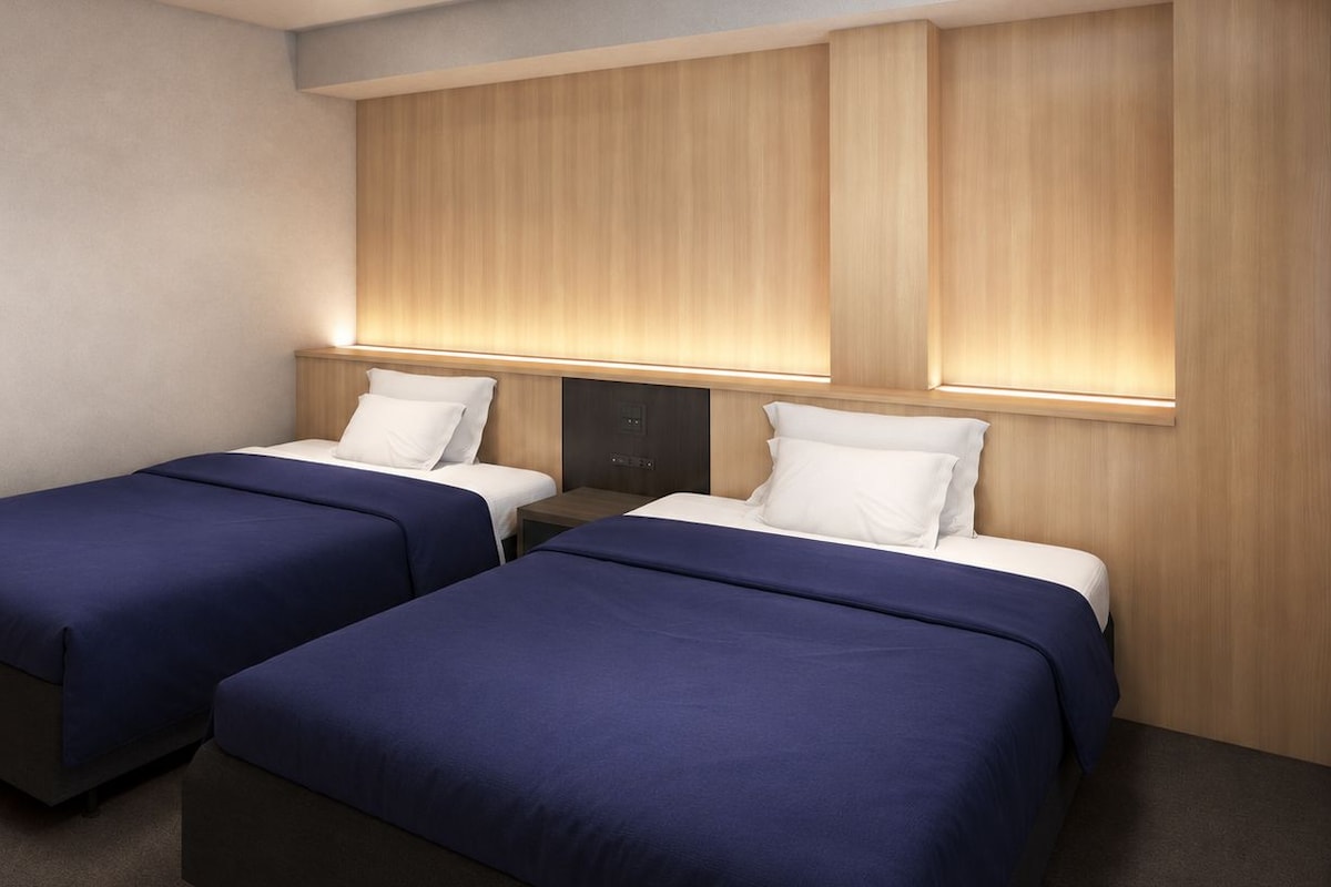 A hotel room with two beds, each with a blue bedspread and white pillows, against a wall with wooden paneling and underglow lighting.