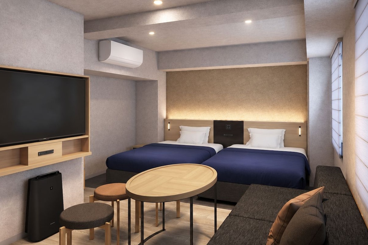 A modern hotel room with a flat-screen TV, two beds, a sofa, a small table, and a wall-mounted air conditioner.