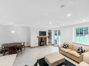 A modern living room with a fireplace, a TV mounted above it, a dining area with a table and chairs, and a large window with a view of greenery. - Coach House - UK52274 (Helmsley)